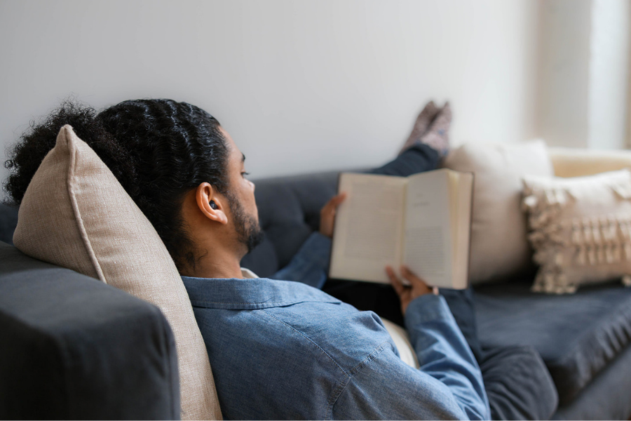 Man reading a book wearing earplugs on a couch in a cozy living room.