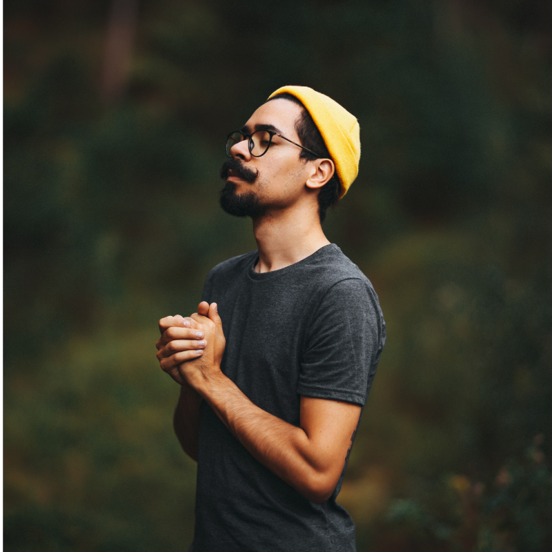 Man wearing a yellow beanie and dark t-shirt with hands clasped in front of him against a blurred natural background